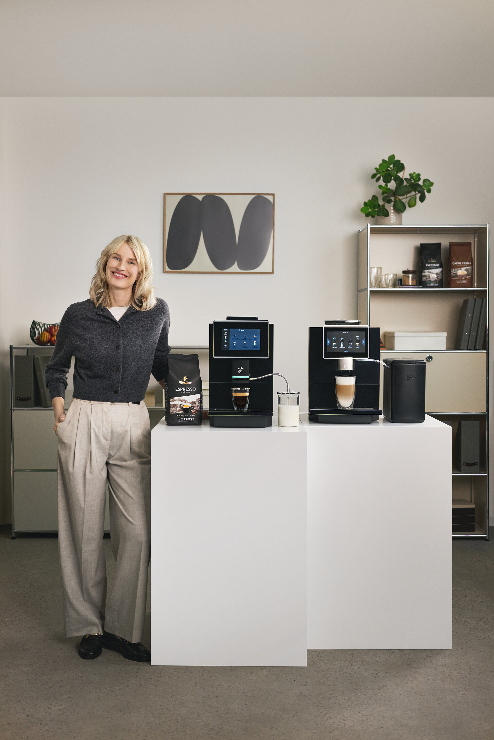 Eine lächelnde Frau steht neben zwei Tchibo Kaffeemaschinen auf weißen Podesten. Auf dem linken Podest steht eine Packung Tchibo Espresso. Eine Tasse Espresso steht unter der linken Maschine.