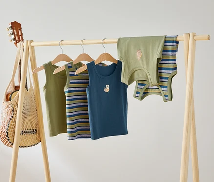 Auf einer Holzstange hängen vier Kinder Tanktops in Grün, gestreift und Blau mit Tiermotiven. Eine Gitarre hängt in einer Netztasche an der Stange.