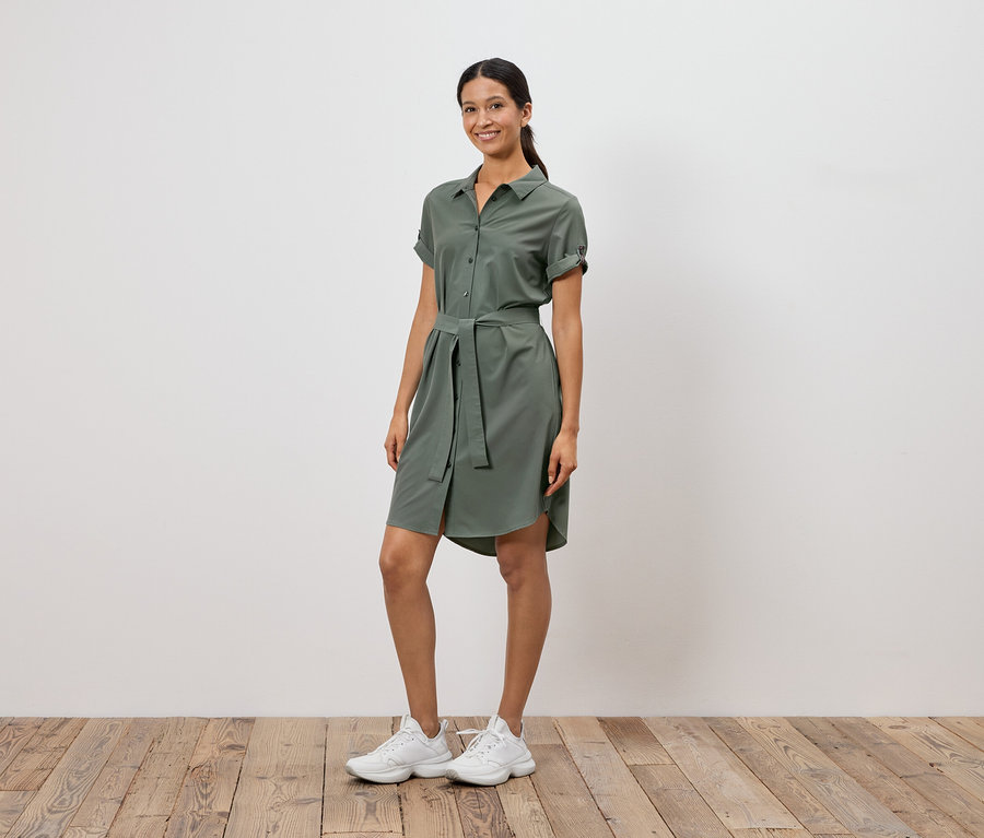 Frau steht vor einer weißen Wand und trägt ein olivgrünes Hemdkleid mit Gürtel und weiße Turnschuhe.