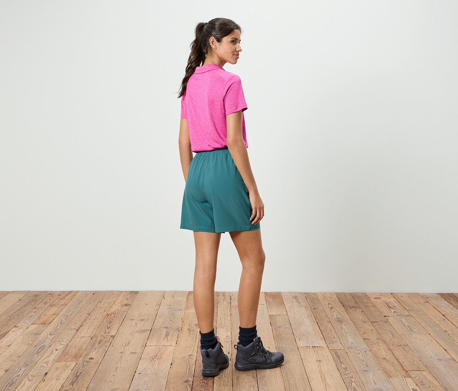 Eine Frau steht mit dem Rücken zur Kamera, sie trägt ein pinkes Poloshirt und grüne Shorts.