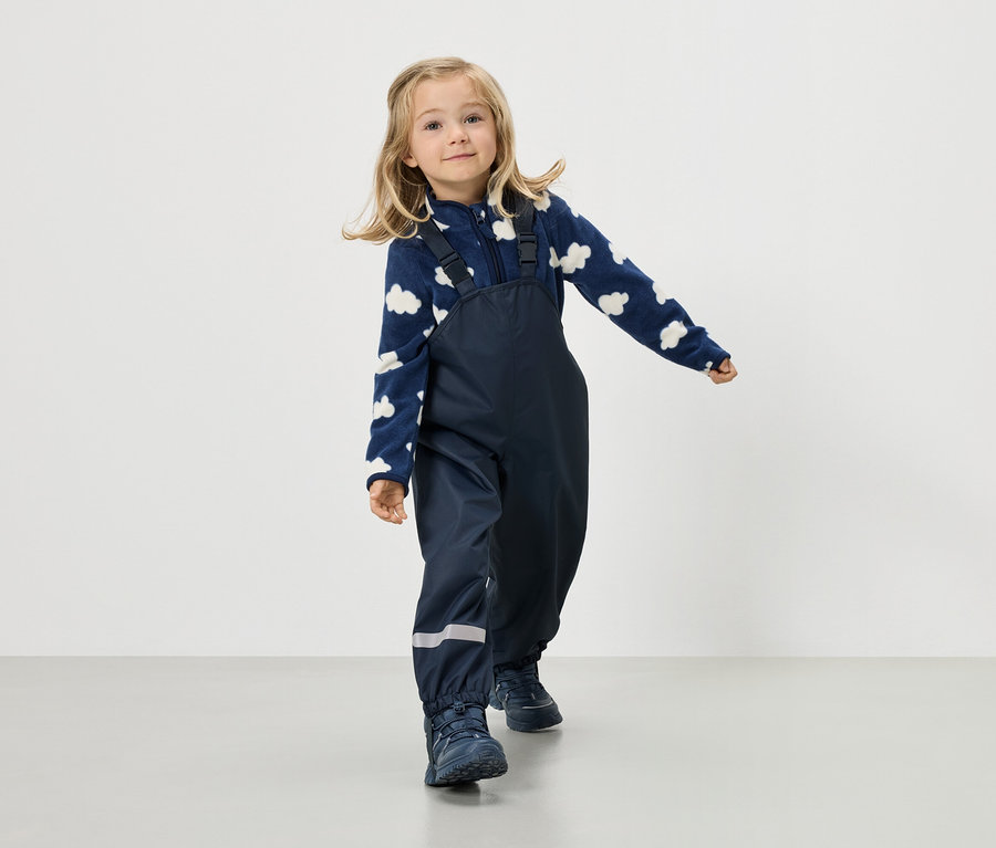 Ein blondes Mädchen trägt eine marineblaue Fleecejacke mit Wolkenmuster und eine marineblaue Hose mit Hosenträgern.
