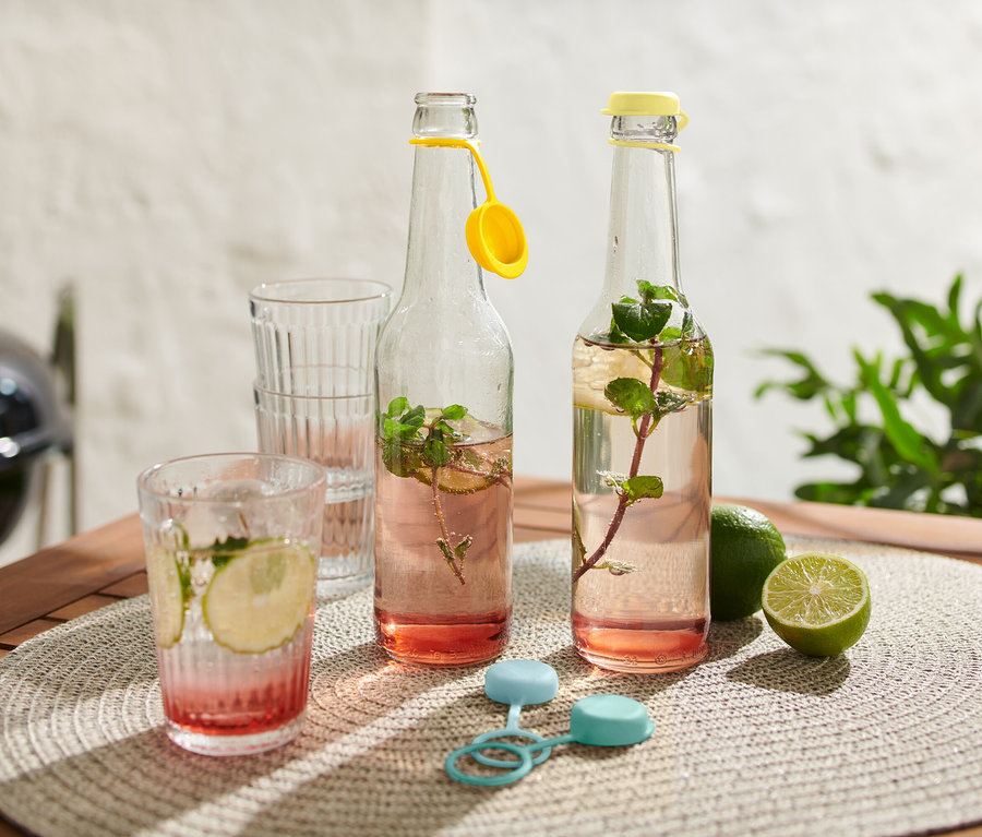 Zwei Wasserflaschen mit gelben Verschlüssen und Minze stehen auf einem Tisch neben Gläsern und Limetten.