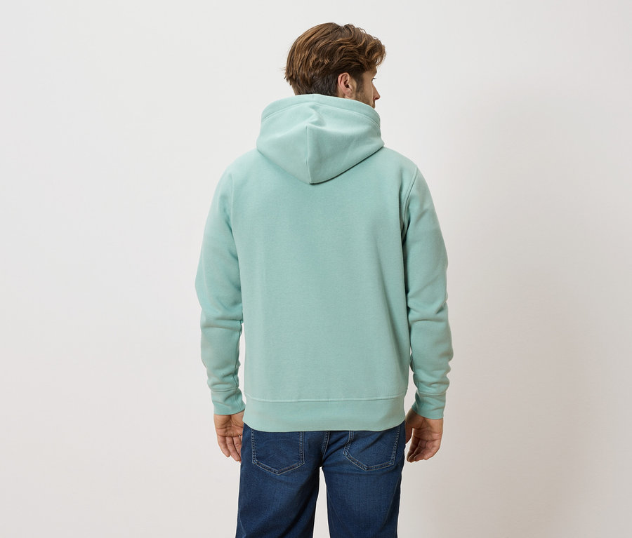 Ein Mann mit braunen Haaren trägt einen mintgrünen Hoodie und blaue Jeans.