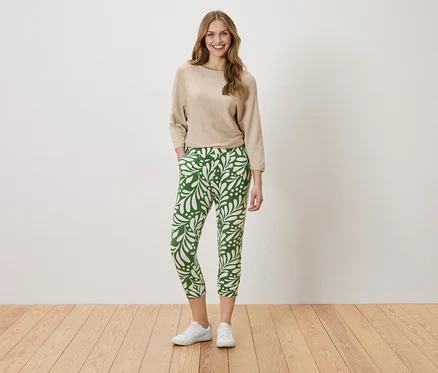 Frau steht mit den Händen in den Taschen vor einer Wand. Sie trägt einen beigefarbenen Pullover und eine grüne Hose mit weißem Blattmuster.