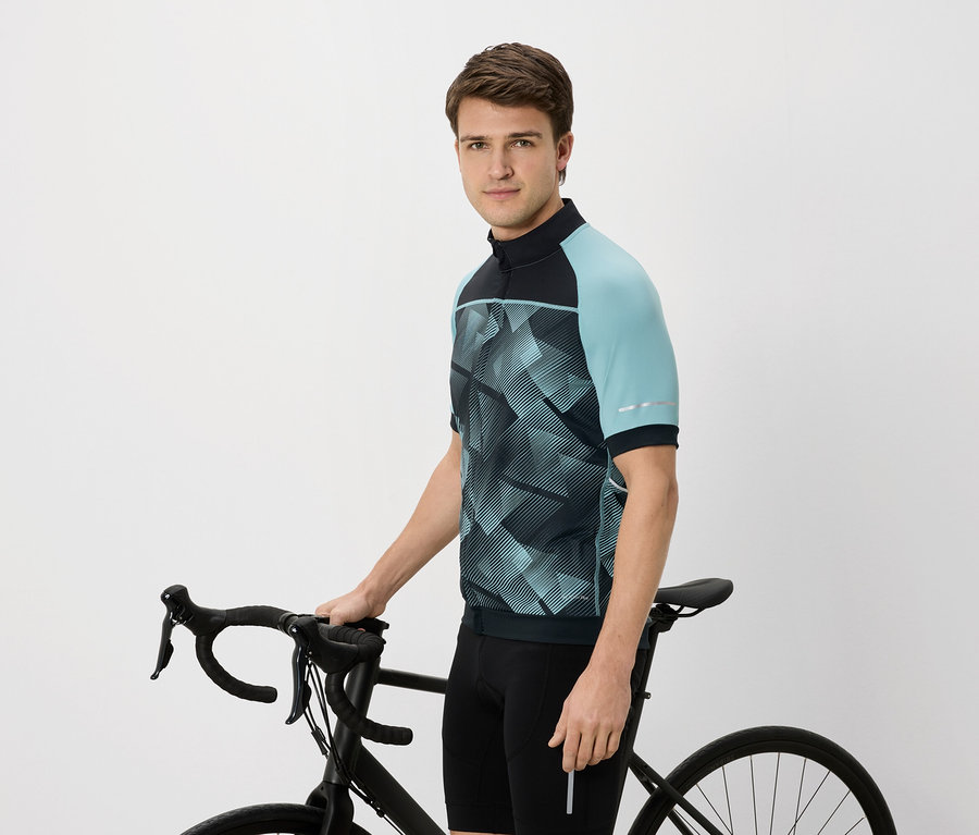 Mann im Radtrikot mit blaugrünen Ärmeln, der ein schwarzes Fahrrad hält.