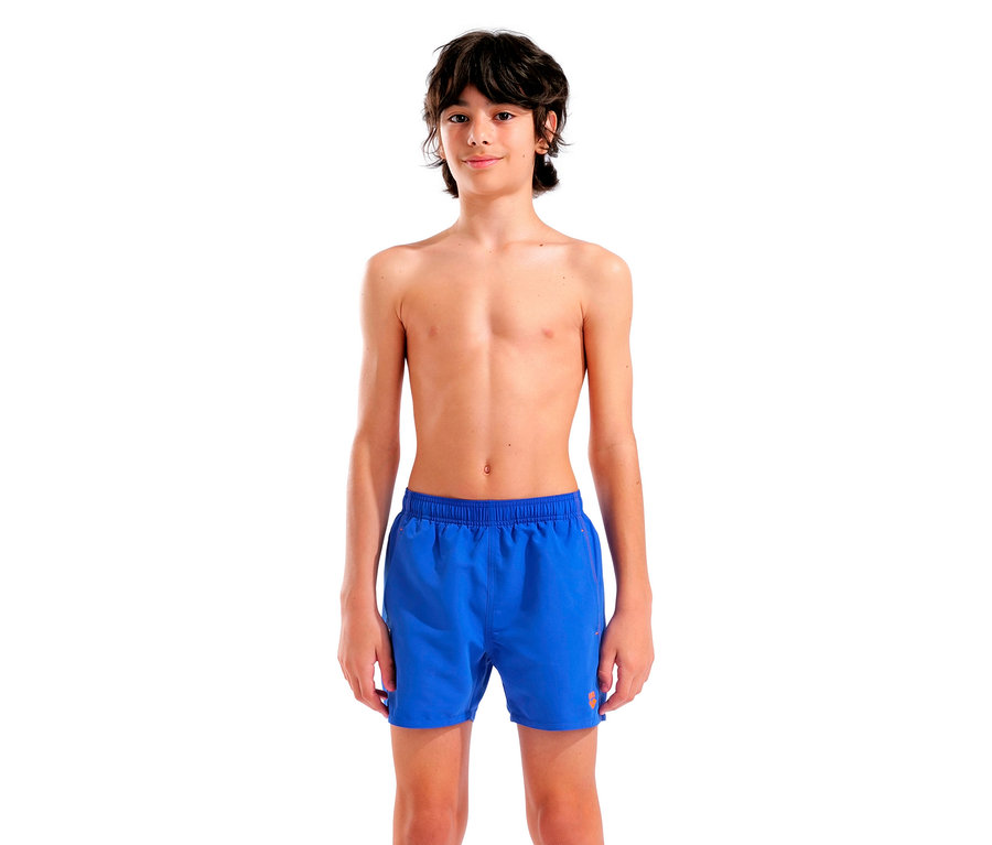 Ein barfüßiger, schlanker Teenager mit dunklen, gewellten Haaren steht vor einem weißen Hintergrund und trägt blaue Badehosen.