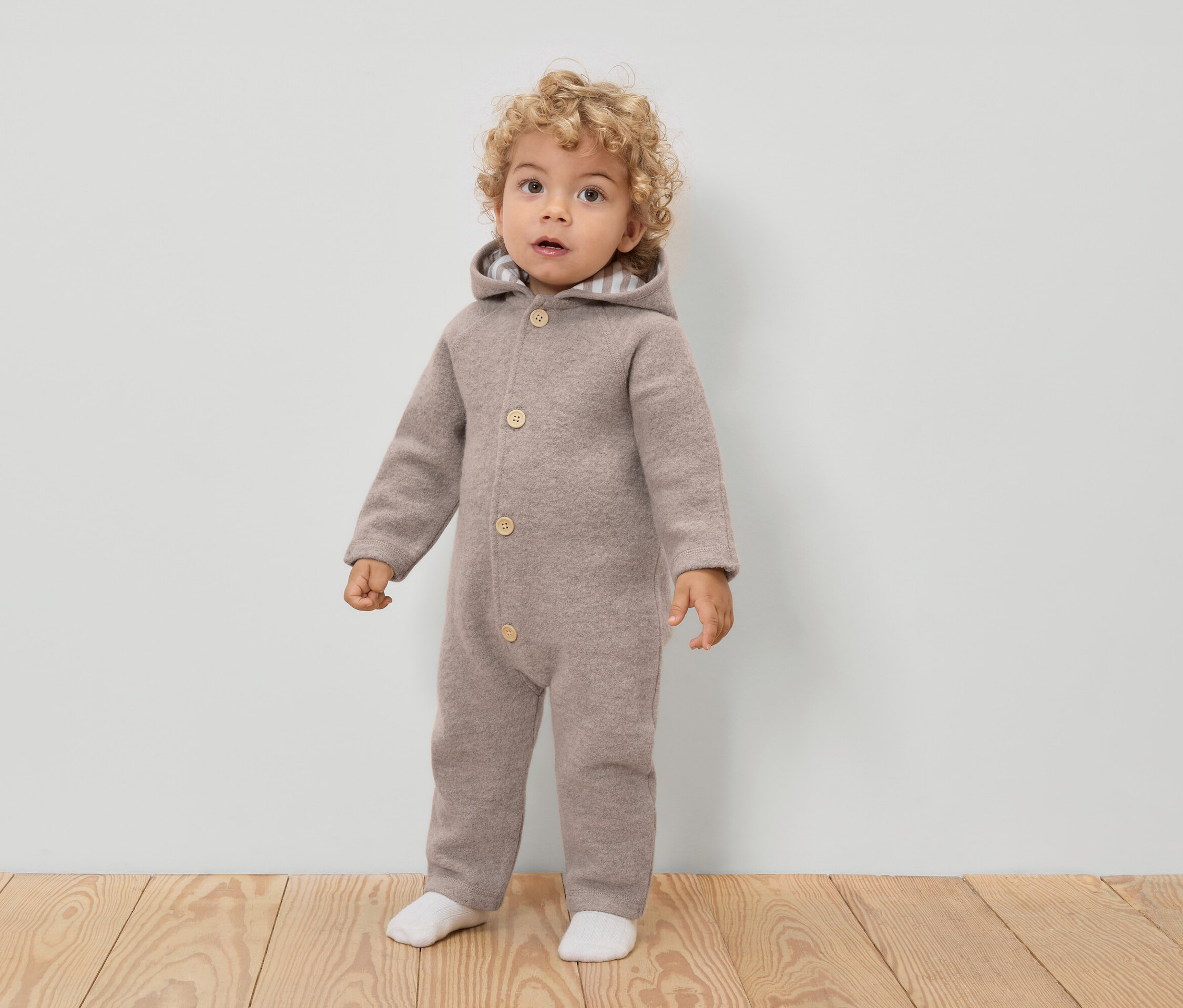 Ein Kleinkind mit lockigen Haaren trägt einen hellbraunen Woll-Overall mit Kapuze und Knöpfen, dazu weiße Socken.