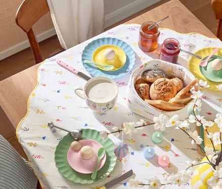 Esstisch mit Gedeck, inklusive Tellern mit Eiern, Tassen, Besteck, Brotkorb, Marmeladengläsern, Osterdekoration und einem blumigen Tischläufer.