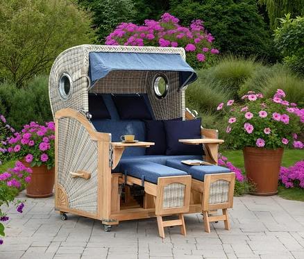 Ein geflochtener Strandkorb mit blauem Stoffdach und blauen Kissen steht auf einer Terrasse, umgeben von rosa blühenden Pflanzen.