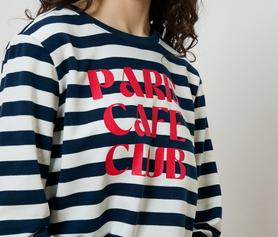 Frau trägt ein blau-weiß gestreiftes Sweatshirt mit der Aufschrift "Paris Cafe Club" in Rot.
