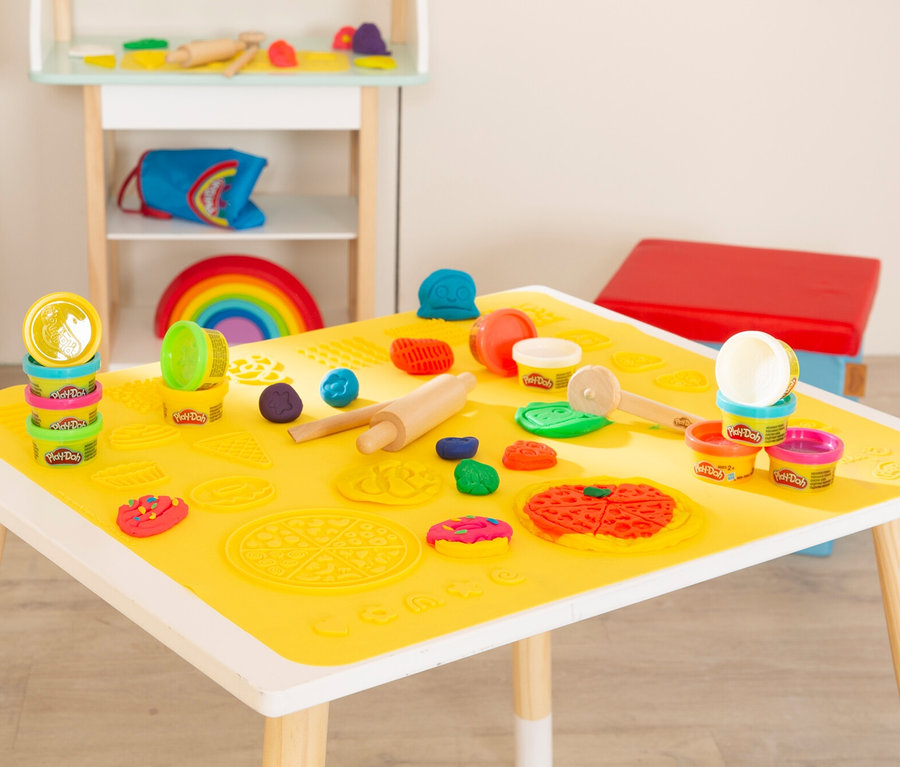 Auf einem Tisch mit gelber Matte liegen Play-Doh-Dosen, Förmchen und Werkzeuge. Im Hintergrund stehen ein Regal und ein Hocker.