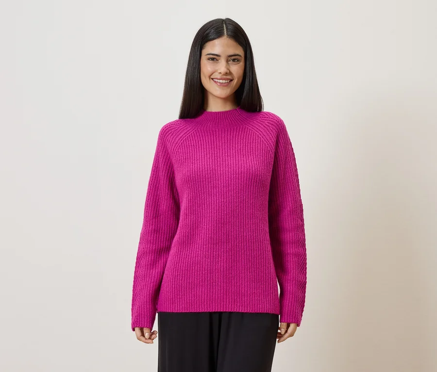 Frau mit dunklen Haaren trägt einen pinken Strickpullover und schwarze Hose vor einem hellen Hintergrund.