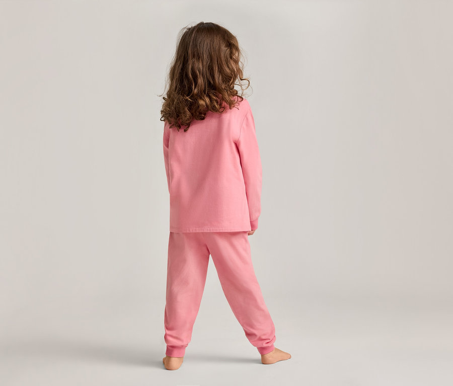 Ein Kind mit lockigem Haar steht in einem rosa Pyjama-Set mit Blick auf die Rückseite vor einem hellgrauen Hintergrund.