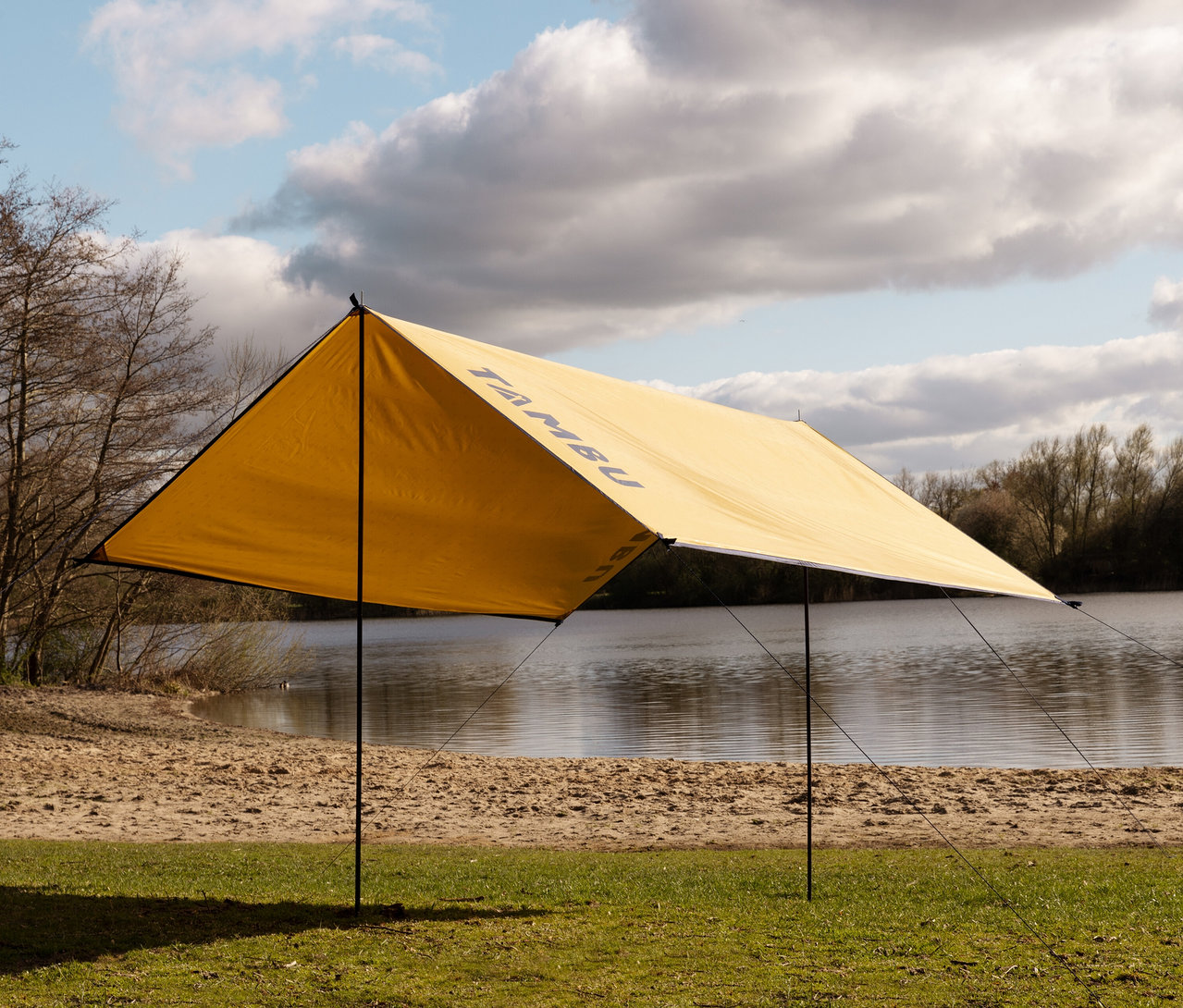 Ein gelbes Tambu »CHADA« Tarp/Sonnensegel steht am Ufer eines Sees.