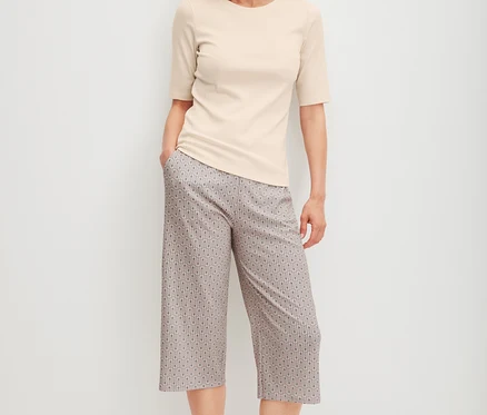 Eine lächelnde Frau steht in einem hellen, kurzärmeligen Oberteil und einer gemusterten CALIDA »Favorites Arts« 3/4-Hose mit den Händen in den Taschen.