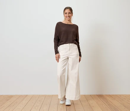 Eine Frau steht vor einer Wand und trägt einen braunen Pullover, eine weite, helle Hose und weiße Schuhe.