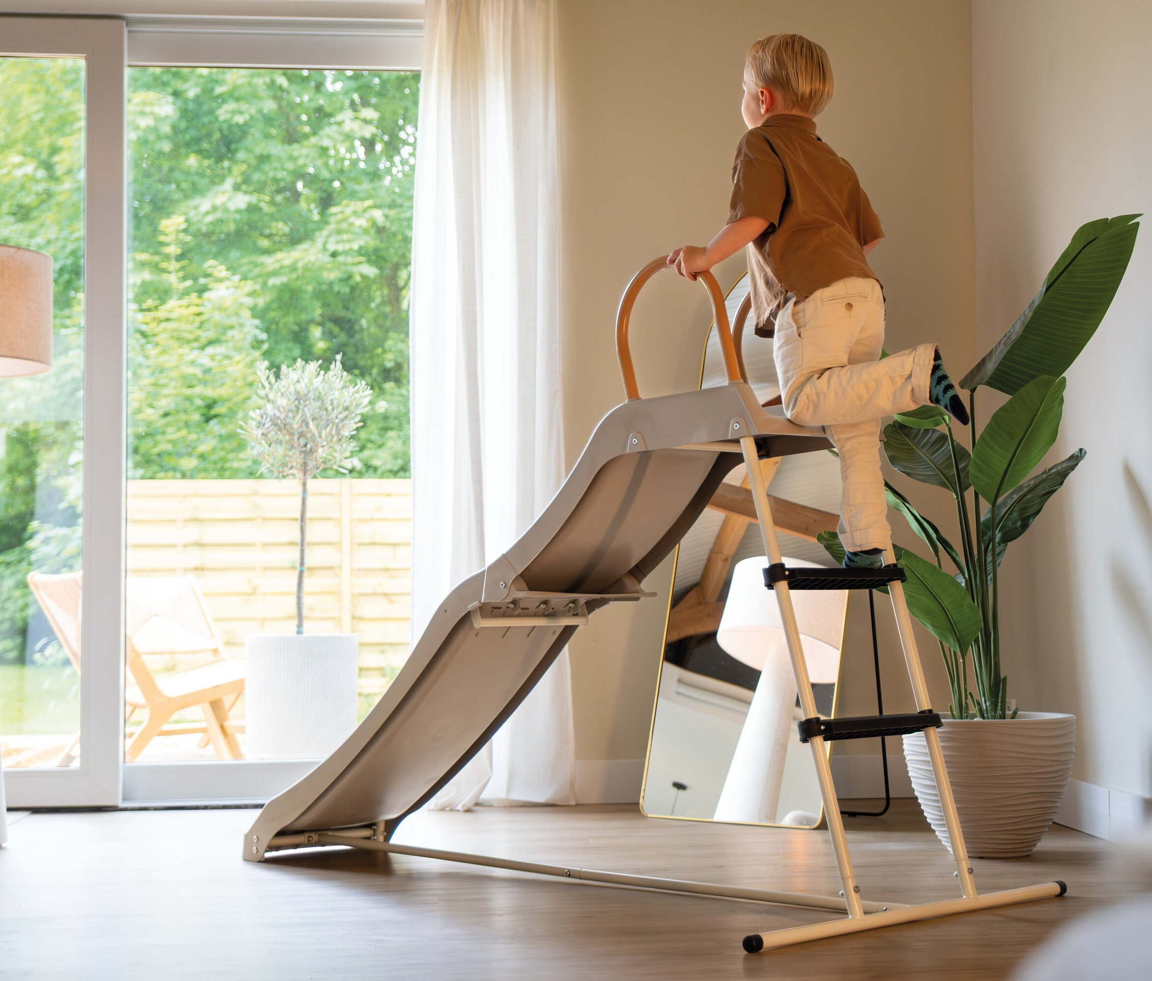 Junge klettert auf eine graue Kinderrutsche mit Leiter in einem hellen Raum. Eine Zimmerpflanze und ein Spiegel stehen daneben.