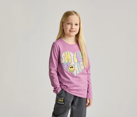 Mädchen mit langen blonden Haaren, trägt ein lila Langarmshirt mit einem bunten Herzaufdruck und graue Jogginghose.