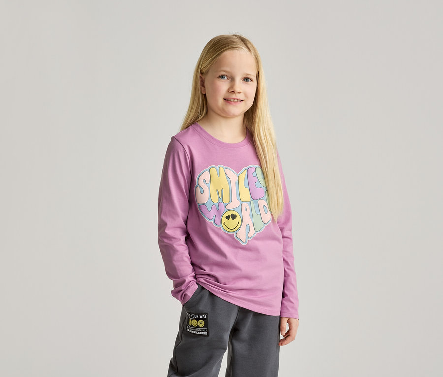 Mädchen mit langen blonden Haaren, trägt ein lila Langarmshirt mit einem bunten Herzaufdruck und graue Jogginghose.