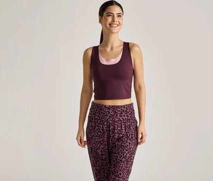 Frau mit Pferdeschwanz trägt bordeauxfarbenes Tanktop über einem rosa Top und einer Hose mit Leopardenmuster vor einem hellen Hintergrund.