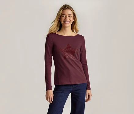 Lächelnde Frau mit langen blonden Haaren trägt ein weinrotes Langarmshirt mit strukturiertem Sternmotiv und dunkelblauer Jeans.