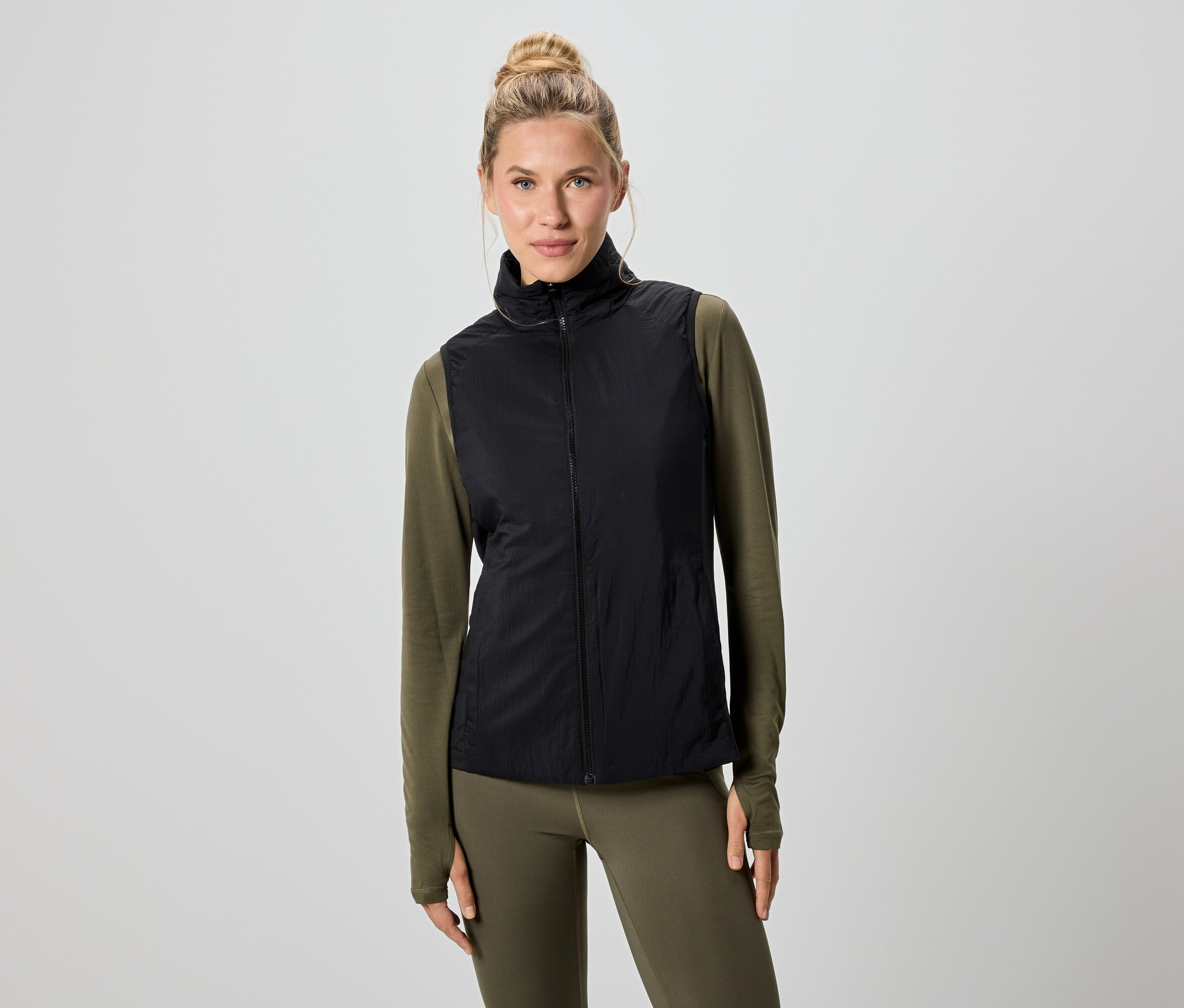 Eine Frau mit blonden Haaren trägt eine schwarze Weste über einem grünen Langarmshirt und Leggings.