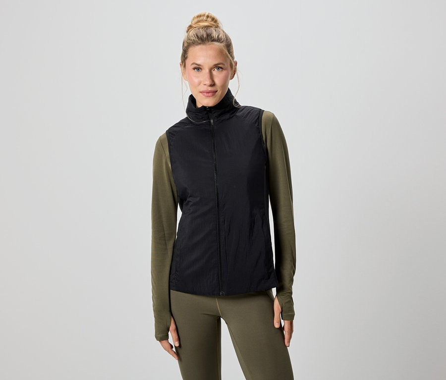 Eine Frau mit blonden Haaren trägt eine schwarze Weste über einem grünen Langarmshirt und Leggings.