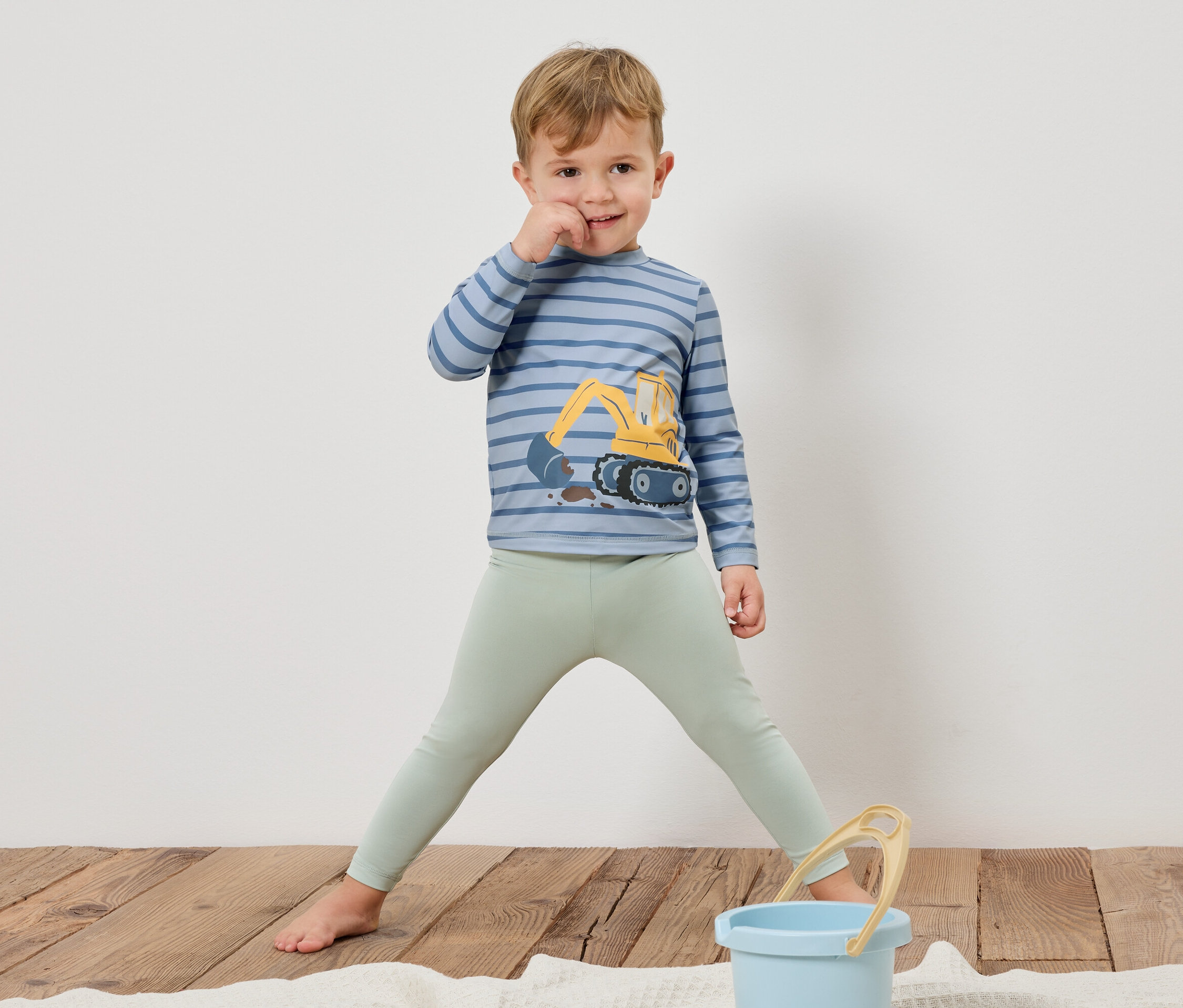 Ein Junge steht barfuß auf einem Holzboden, trägt ein gestreiftes Langarmshirt mit einem Bagger darauf und hellgrüne Leggings. Ein blauer Eimer steht vor ihm.