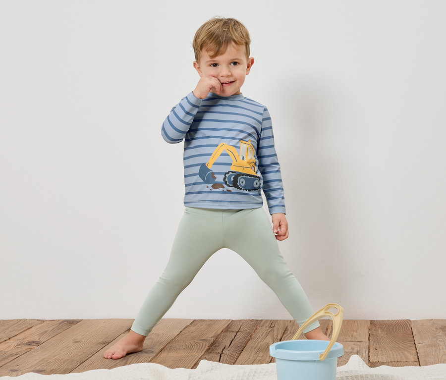 Ein Junge steht barfuß auf einem Holzboden, trägt ein gestreiftes Langarmshirt mit einem Bagger darauf und hellgrüne Leggings. Ein blauer Eimer steht vor ihm.