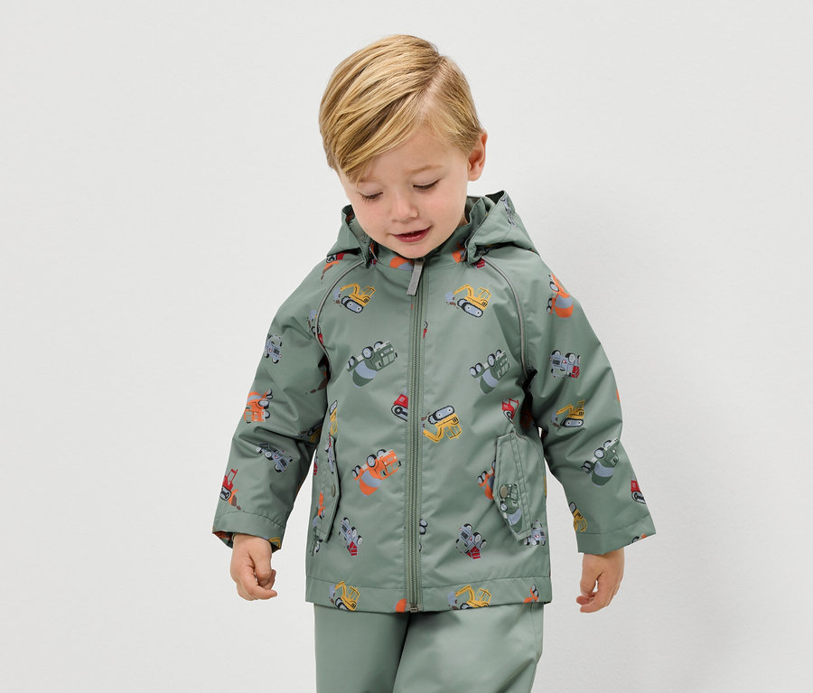Ein blonder Junge trägt eine grüne Regenjacke mit Bagger-Motiv und eine passende Regenhose.
