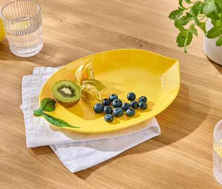 Ein gelber Teller mit Blaubeeren, Kiwi und Physalis liegt auf einem weißen Tuch. Daneben stehen ein Glas Wasser mit Zitrone und eine Zitrone.