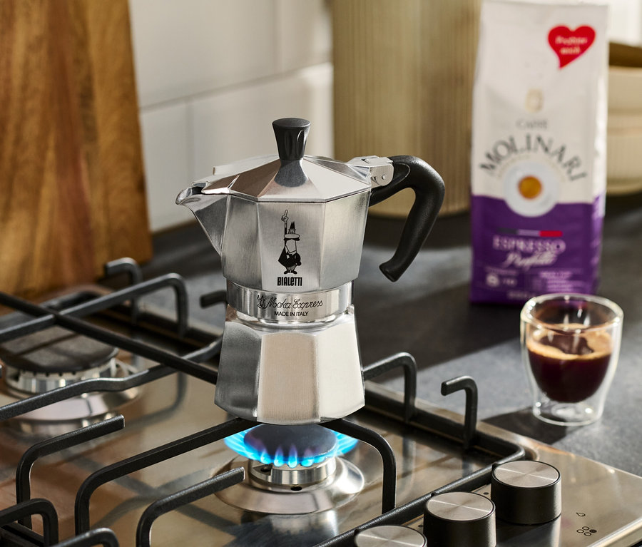 Ein silberner Espressokocher steht auf einem Gasherd mit blauer Flamme, daneben eine Tasse Espresso und eine Packung Molinari-Kaffee.