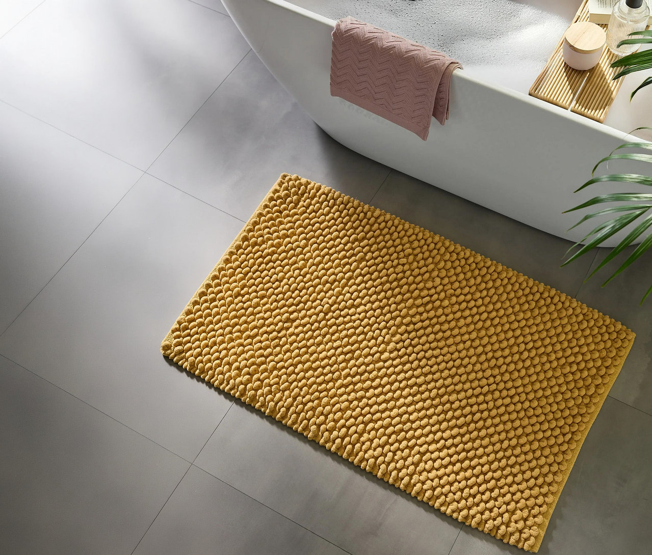 Gelbe Noppen-Badematte auf grauem Fliesenboden neben einer weißen Badewanne mit Schaum, einem rosa Handtuch und einem Holzbadebrett mit Badeartikeln.
