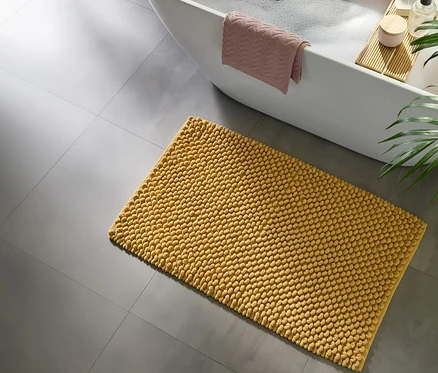 Gelbe Noppen-Badematte auf grauem Fliesenboden neben einer weißen Badewanne mit Schaum, einem rosa Handtuch und einem Holzbadebrett mit Badeartikeln.