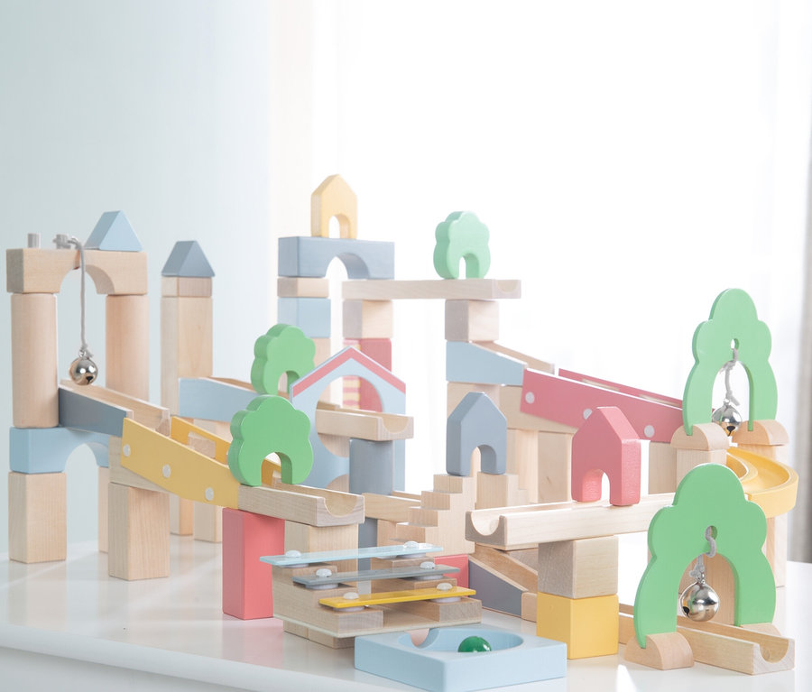 Holzbausteine-Kugelbahn in Pastellfarben, mit Rampen, Bögen, Häusern, Bäumen, Glocken und einem Xylophon auf einem weißen Tisch.