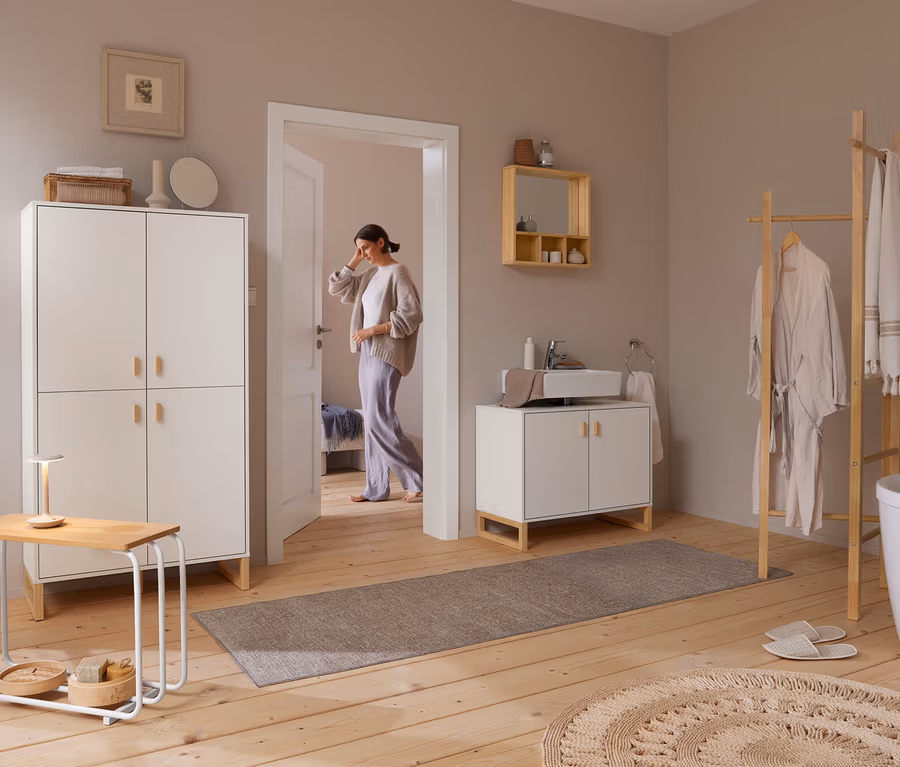 Eine Frau betritt ein helles Badezimmer mit Holzboden. Rechts steht eine Kleiderleiter mit Bademantel und Handtüchern neben einer weißen Badewanne. Über einem weißen Waschtisch mit Waschbecken hängt ein Spiegel aus massivem Bambus mit Ablagen. Links steht ein hoher weißer Schrank neben einem kleinen Tisch mit Lampe.