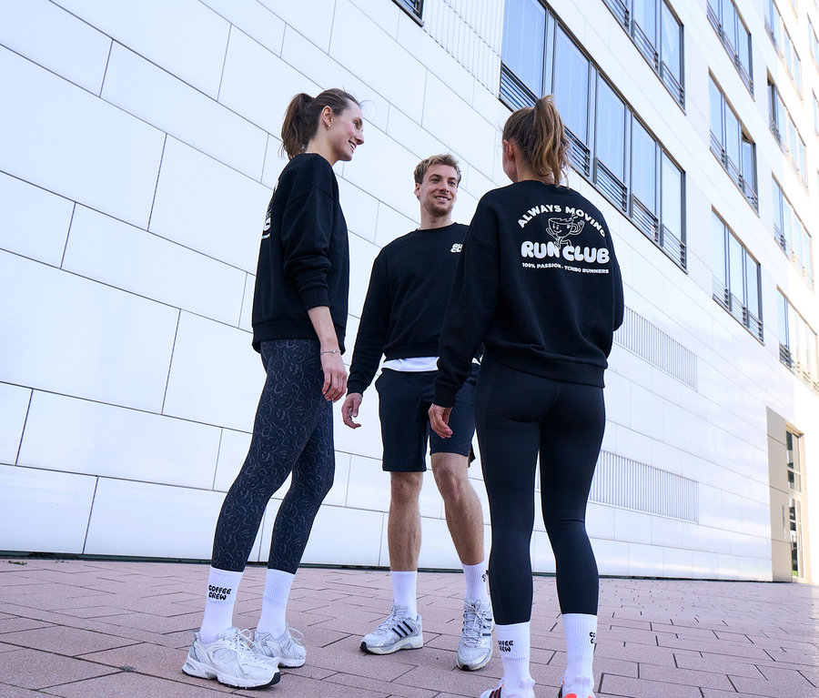 Drei Personen, zwei Frauen und ein Mann, stehen lächelnd und unterhalten sich. Alle tragen schwarze Sweatshirts und weiße Unisex Coffee Crew Tennissocken. Der Mann trägt schwarze Sportshorts. Eine Frau trägt grau-schwarze Sportleggings »ActivePower«, die andere schwarze Sportleggings »ActivePower«.