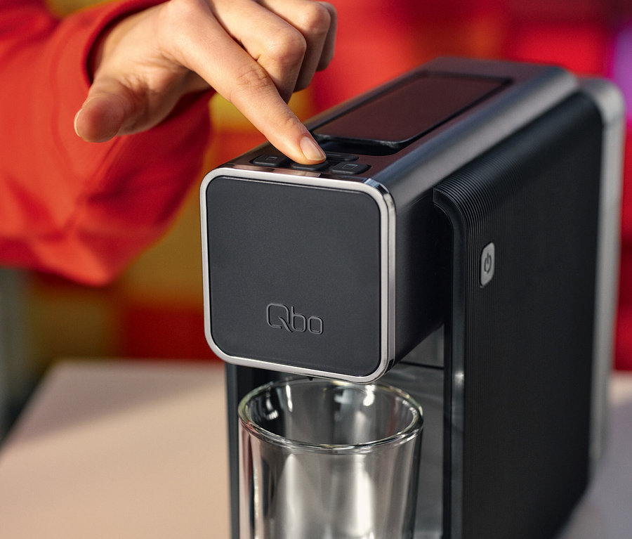 Eine Person drückt einen Knopf auf einer schwarzen Qbo-Kaffeemaschine. Darunter steht ein leeres Glas.