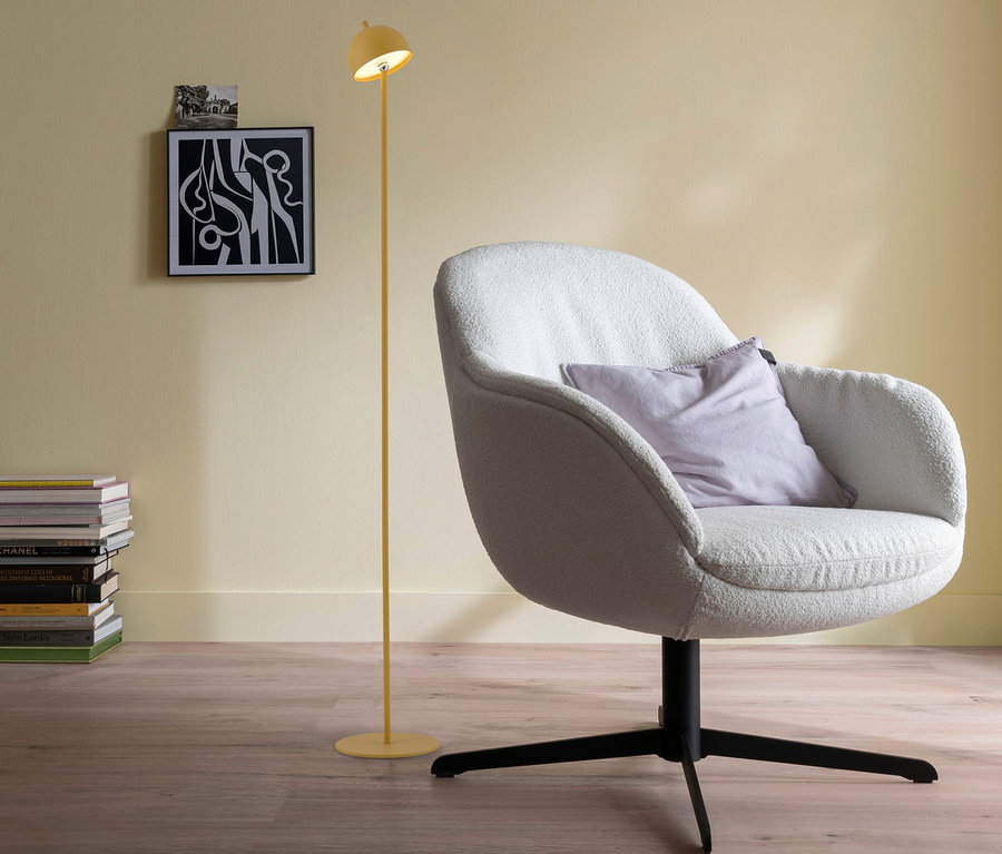 Gelbe SCHÖNER WOHNEN-Kollektion Akku-Stehleuchte »Bellis« steht neben einem hellen Sessel mit lila Kissen, Bücherstapel und Bildern an der Wand.