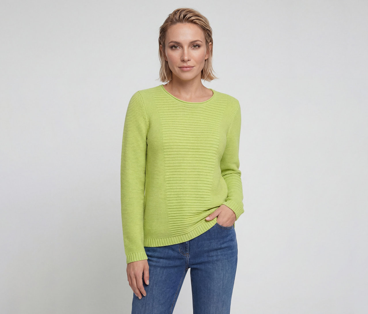 Frau mit kurzen blonden Haaren trägt einen limettengrünen Rippstrickpullover und blaue Jeans, eine Hand in der Tasche, blickt in die Kamera.