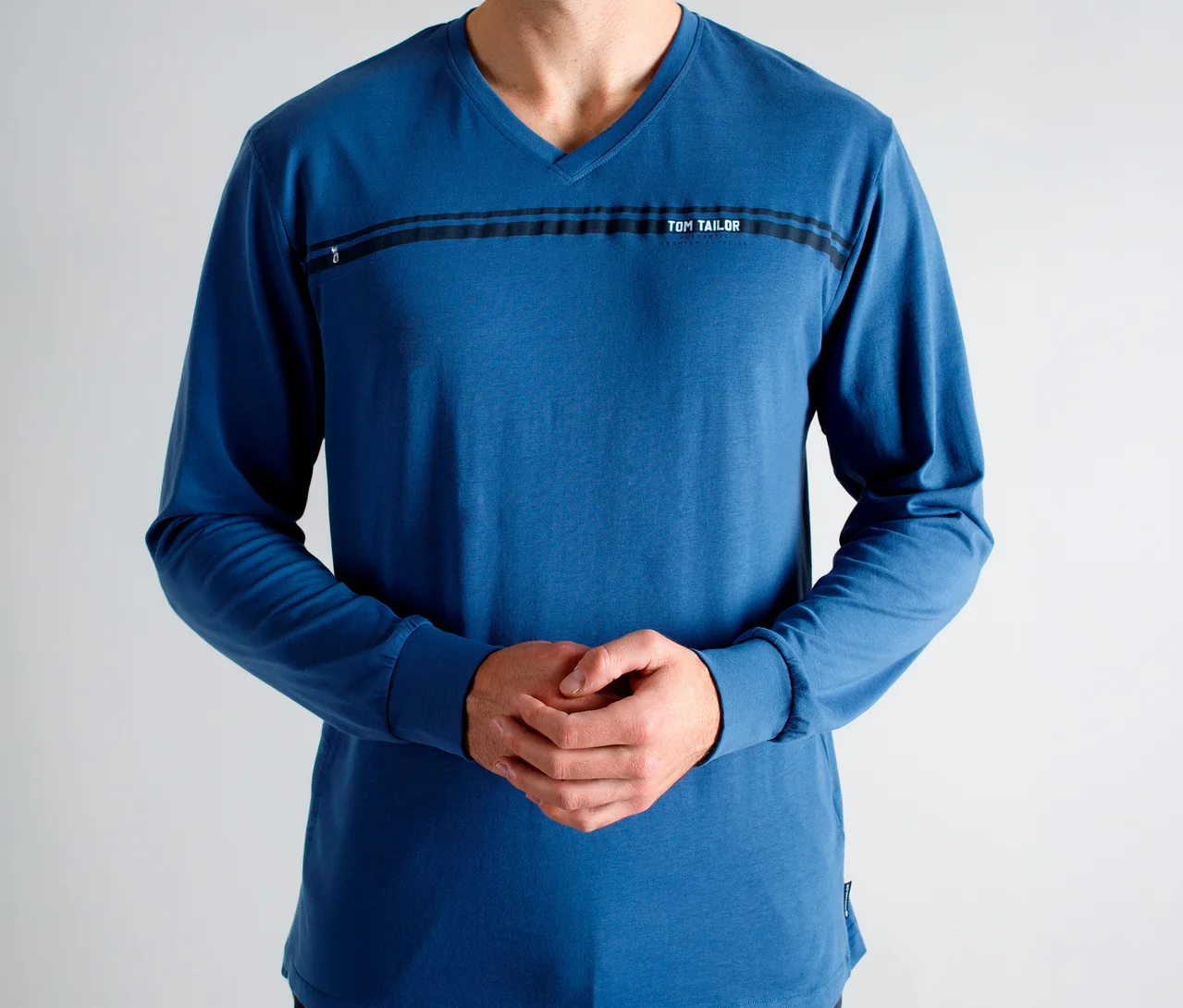Mann in einem blauen Langarm-V-Ausschnitt-Shirt mit schwarzen Streifen und „TOM TAILOR“-Schriftzug auf der Brust, Hände gefaltet.
