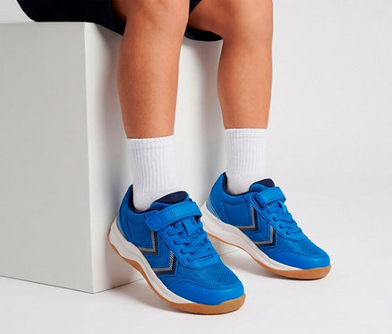 Beine und Füße einer Person mit blauen Turnschuhen mit Klettverschluss und weißen Socken, die auf einem weißen Block sitzt.