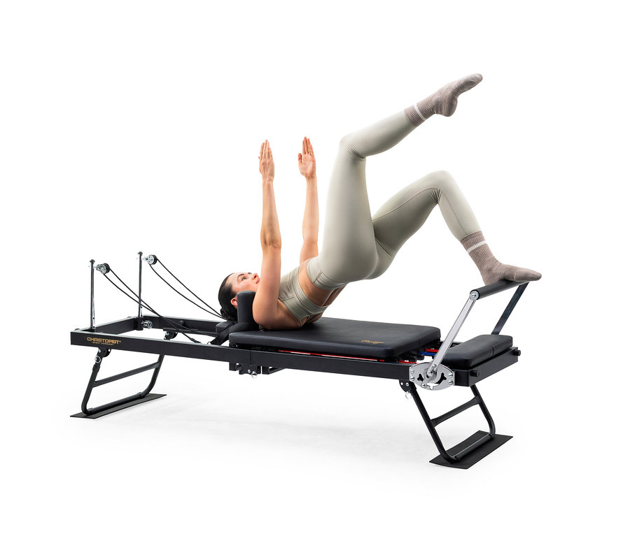 Frau in hellgrüner Sportkleidung macht Pilates auf einem schwarzen Reformer-Gerät, liegt auf dem Rücken mit angehobener Hüfte, erhobenen Armen und Füßen auf der Fußstange.