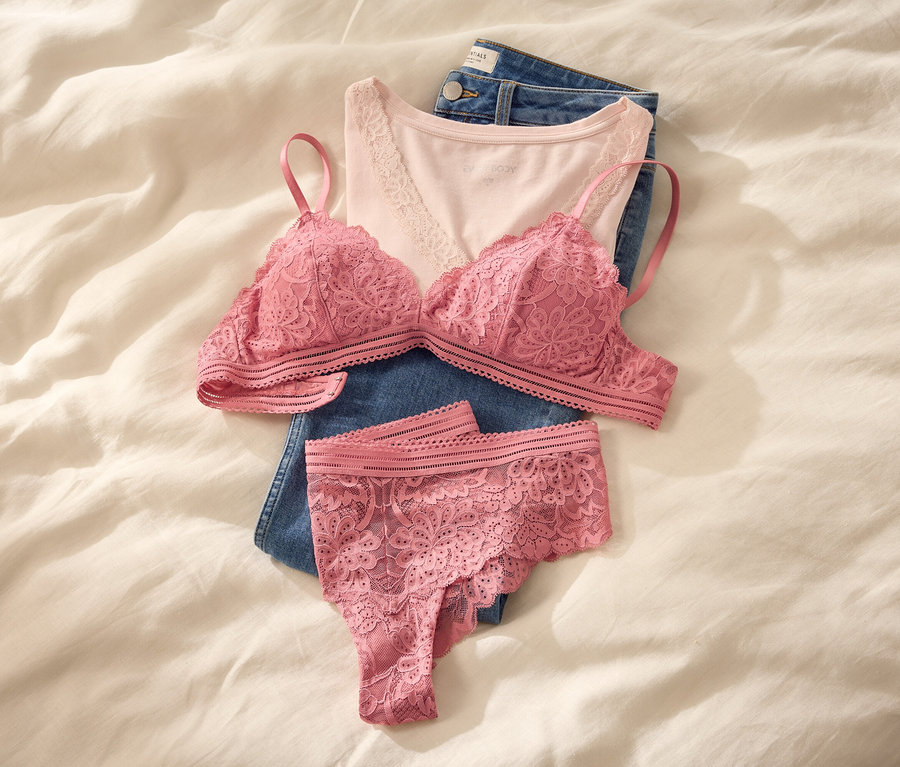 Ein pinker Spitzen-Bralette und passender Tanga liegen auf einem hellrosa Camisole und gefalteten blauen Jeans auf einem weißen Bett.