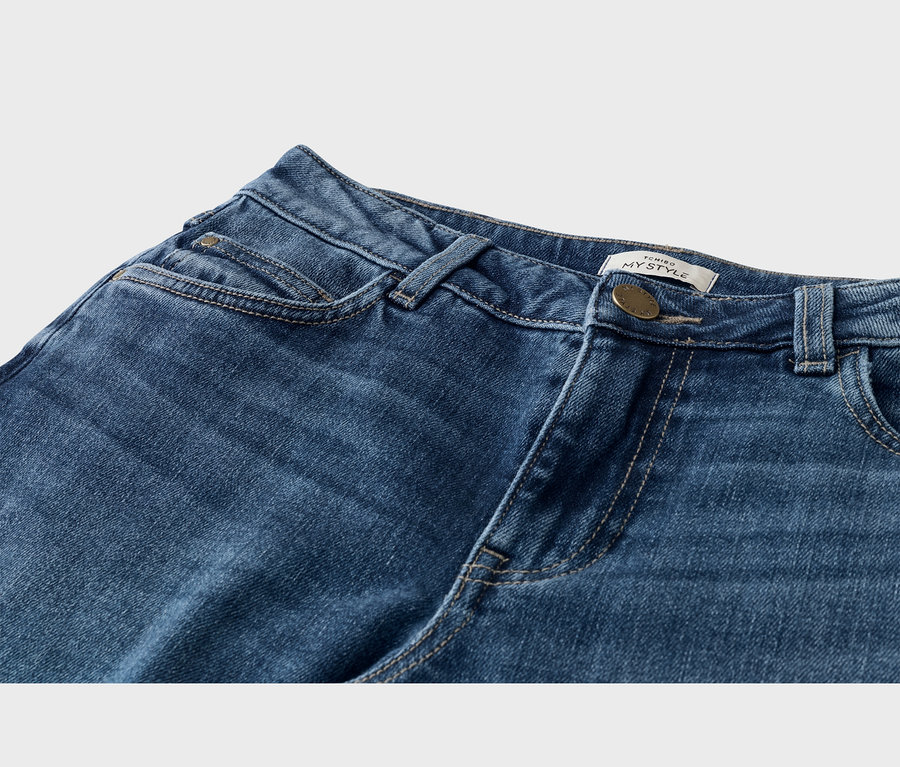 Nahaufnahme einer blauen Jeans mit Bund, Knopf, Gürtelschlaufen und Taschen. Ein weißes Etikett mit „TCHIBO MY STYLE“ ist nahe dem Messingknopf sichtbar.