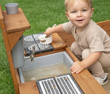 Ein lächelndes Kleinkind in beiger Kleidung spielt mit einer Matschküche aus Holz und schöpft Matsch in eine weiße Schüssel. Die Küche hat ein Waschbecken mit Wasser, einen Wasserhahn und zwei gemusterte Kochplatten.