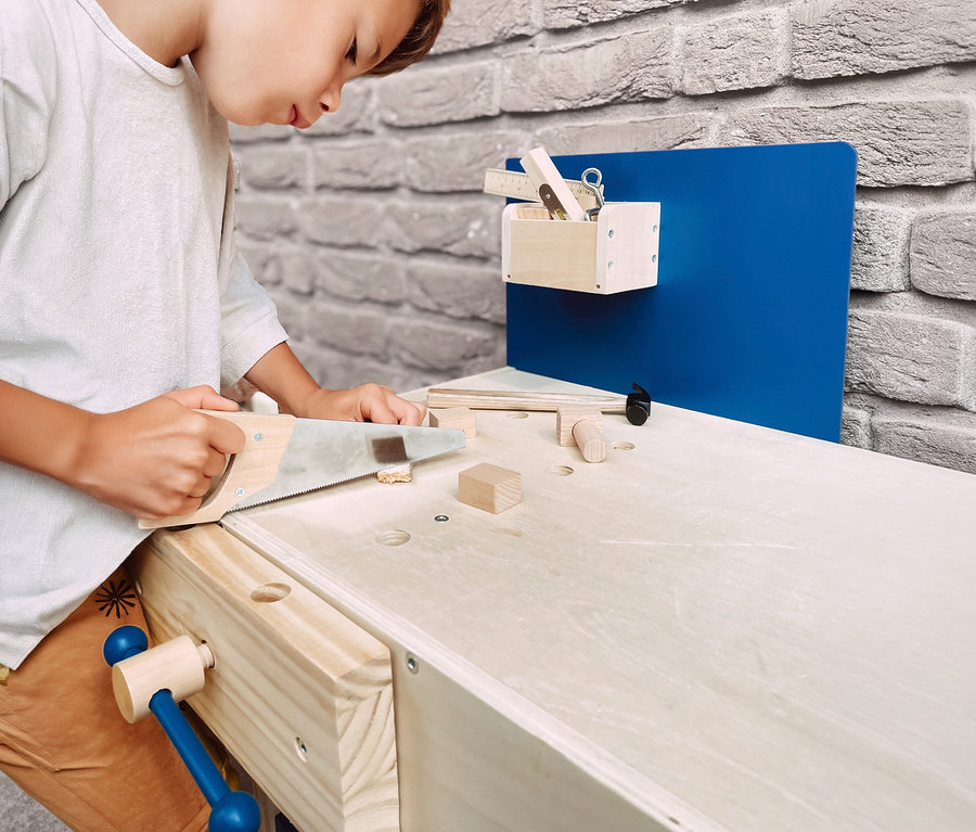 Kind sägt mit einer Spielzeugsäge einen Holzklotz auf einer small foot Kinderwerkbank. Auf der Werkbank liegen weitere Holzspielzeuge und Werkzeuge.