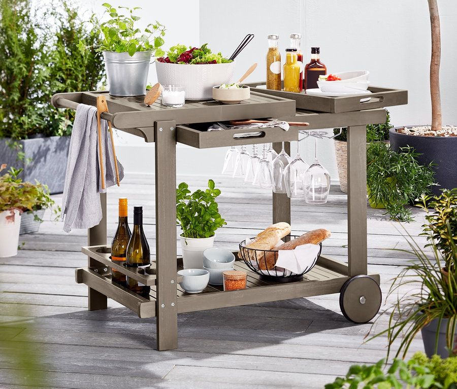 Grauer Barbecue-Trolley mit Salat, Getränken, Gläsern und Brot auf einer Terrasse.