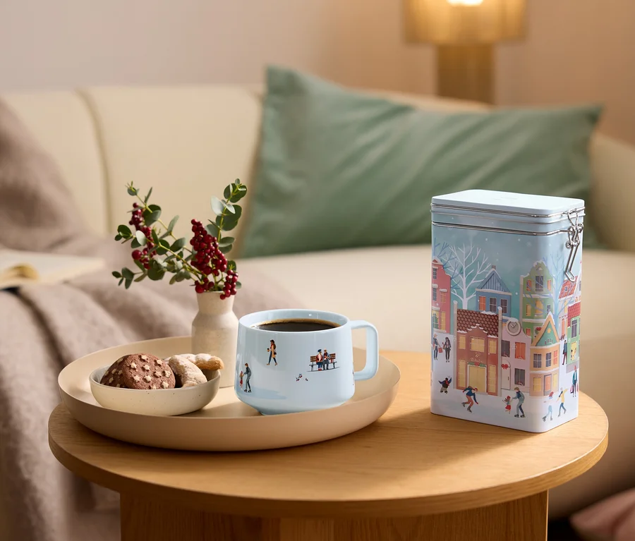 Hellblaue Kaffeetasse mit Winterszene, eine Schale Kekse und eine Vase mit Beeren auf einem Tablett auf einem Holztisch. Eine hellblaue Blechdose mit Winterdorf-Illustration steht daneben, mit einem Sofa im Hintergrund.