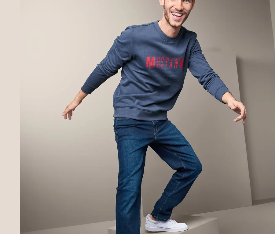 Ein Mann posiert auf einem grauen Würfel in einem blauen Sweatshirt mit rotem Schriftzug und Jeans »Mustang«.